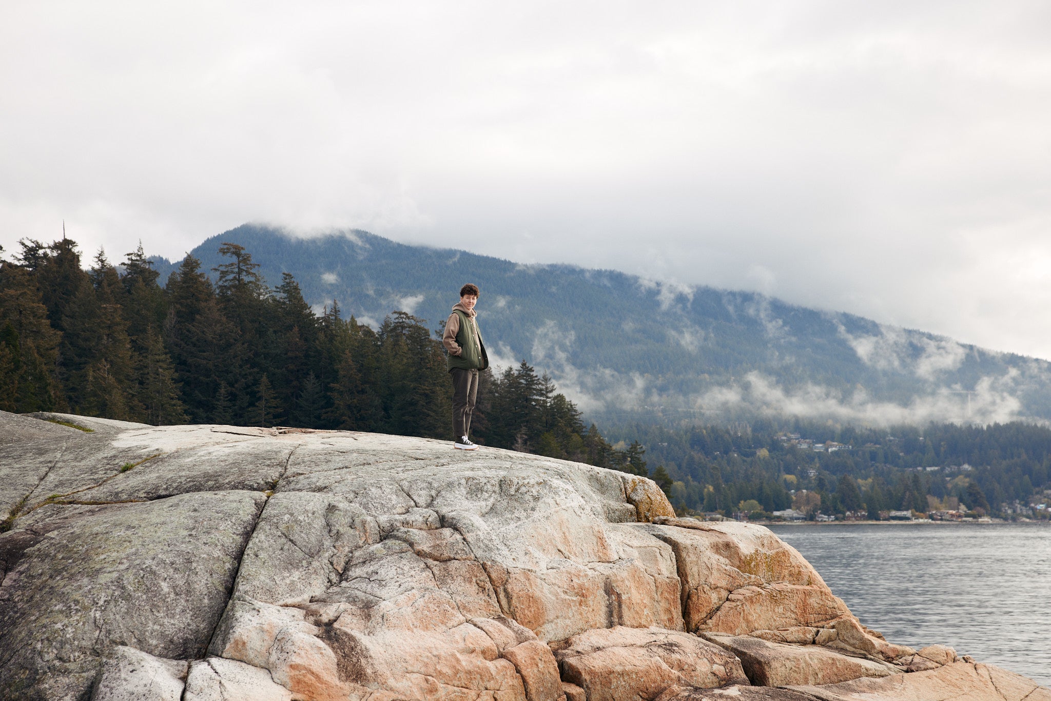 AK MacKellar wears ecologyst apparel while standing on a cliff with the ocean and mountains in the background.